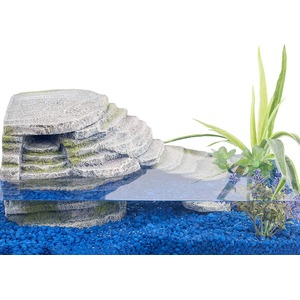 Tiered stone rock formation beside clear water, surrounded by blue gravel and green plants.