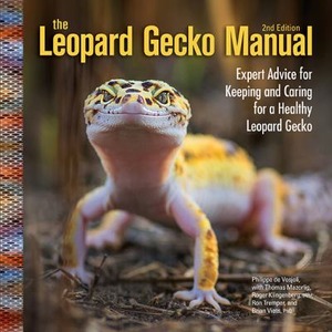 Leopard gecko close-up on soil with a book cover reading “The Leopard Gecko Manual” 2nd Edition.